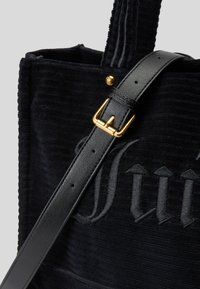 Saco de lona preto em corduroy com uma superfície texturizada, apresentando uma fivela de metal dourado numa alça de couro preto e detalhes bordados.