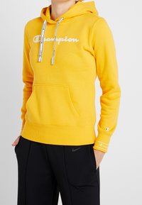Sweat à capuche jaune en tissu doux, arborant le logo "Champion" blanc, cordons de serrage avec accents noirs, et une poche avant. Associé à un pantalon noir.