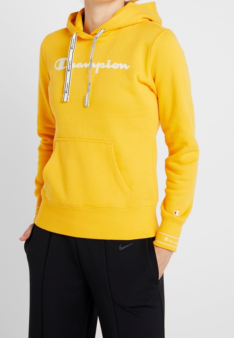 Sweat à capuche jaune en tissu doux, arborant le logo "Champion" blanc, cordons de serrage avec accents noirs, et une poche avant. Associé à un pantalon noir.