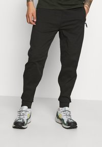 Černé zúžené kalhoty s elastickými manžetami, se bočními kapsami a kapsou na zip. V kombinaci se šedými a zelenými sportovními botami.