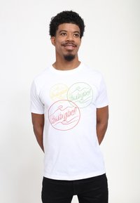 Vit bomull t-shirt med tre cirkulära grafiker i gult, grönt och rött med "Babybel"-text inuti varje cirkel. Standard rundhalsdesign.