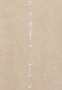 Beiges Baumwollhemd mit vertikaler weißer Schrift "KEY LARG" im Used-Look. Weiche Textur, schlichtes Design, lässiger Stil.