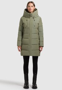 Olīvu zaļš pūkains ceļojumu mētelis ar lielu kapuci, kviltota dizaina, ar auklu aizdari un sānu kabatām. Valkāts pār melnām legingām un zābakiem.