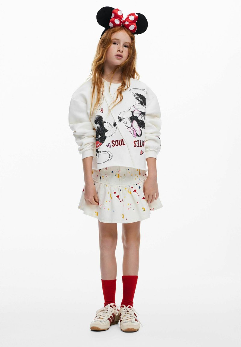 Sudadera blanca con un gráfico de Mickey y Minnie Mouse; combinada con una falda blanca decorada con manchas de colores; calcetines rojos y zapatillas deportivas.