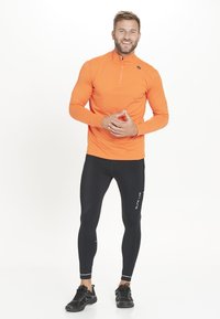 Lys orange langærmet sports shirt med en halv lynlås, parret med tætsiddende sorte leggins og sorte sneakers, hvilket skaber et sporty look.