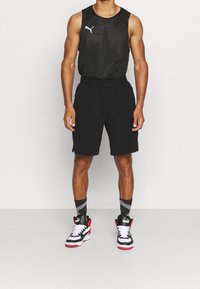 Černý bezrukávový sportovní top s mřížkovou texturou, spárovaný s černými šortkami s kapsami. Nosený s černými a červenými teniskami a vzorovanými ponožkami.