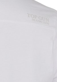 Vit tyg med präglad text som säger "TOP GUN THE FLYING LEGEND" nära den övre kanten.