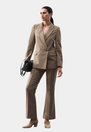 Completo beige su misura con giacca doppiopetto e pantaloni svasati. Presenta una borsa nera e scarpe beige con tacco blocco. Texture del tessuto liscia.