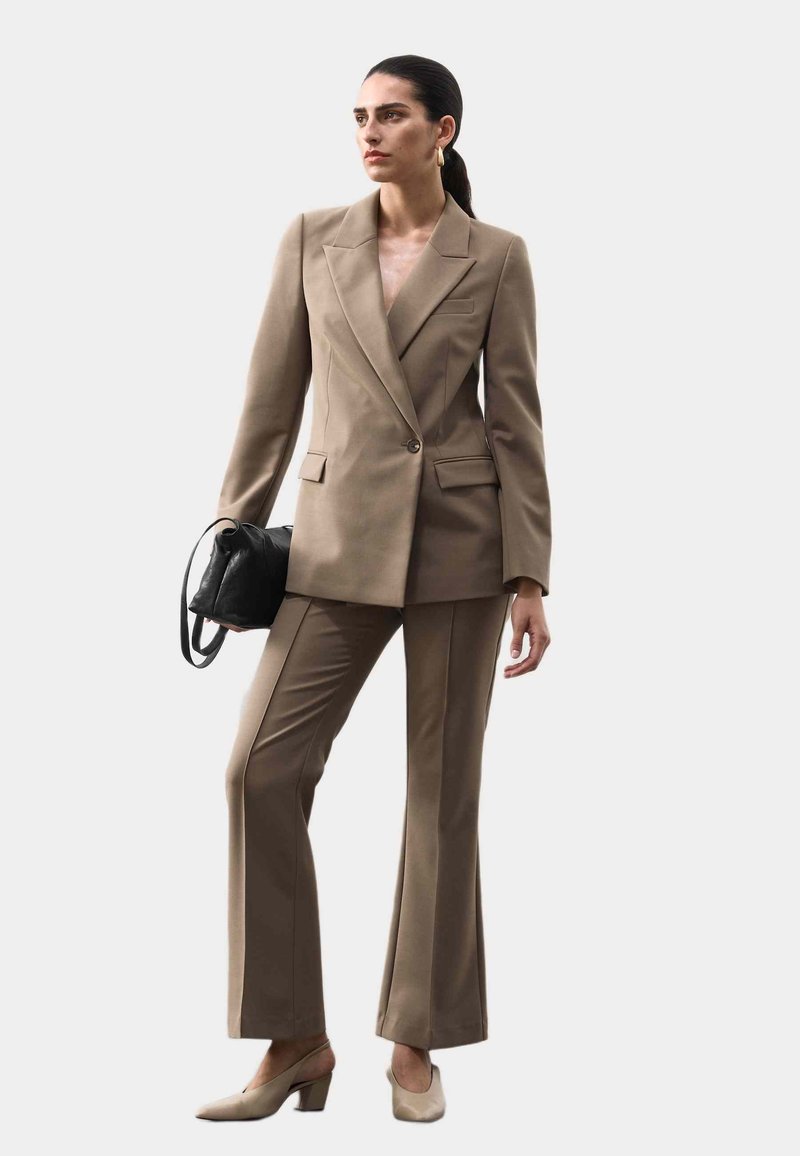 Beige op maat gemaakt pak met een dubbele blazer en flared broek. Beschikt over een zwarte handtas en beige blokhak schoenen. Glad stoffen textuur.