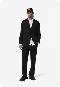 Juodas kostiumo švarkas ir kelnės derinami su balta marškinėliais, papildyti juoda kepure su apskritimu antkyje, dėvimi su sportbačiais.