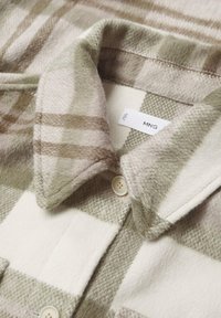 Manteau en laine douce avec un motif à carreaux beige et blanc, col plié et boutons de couleur claire, montrant une petite étiquette de marque à l'intérieur.