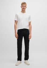 Joven de pie vistiendo una camisa blanca de manga corta lisa, jeans negros y zapatillas blancas frente a un fondo claro y liso.