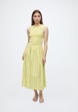 Femme debout portant une robe jaune sans manches avec des rayures verticales bleues et des sandales blanches sur un fond blanc uni.