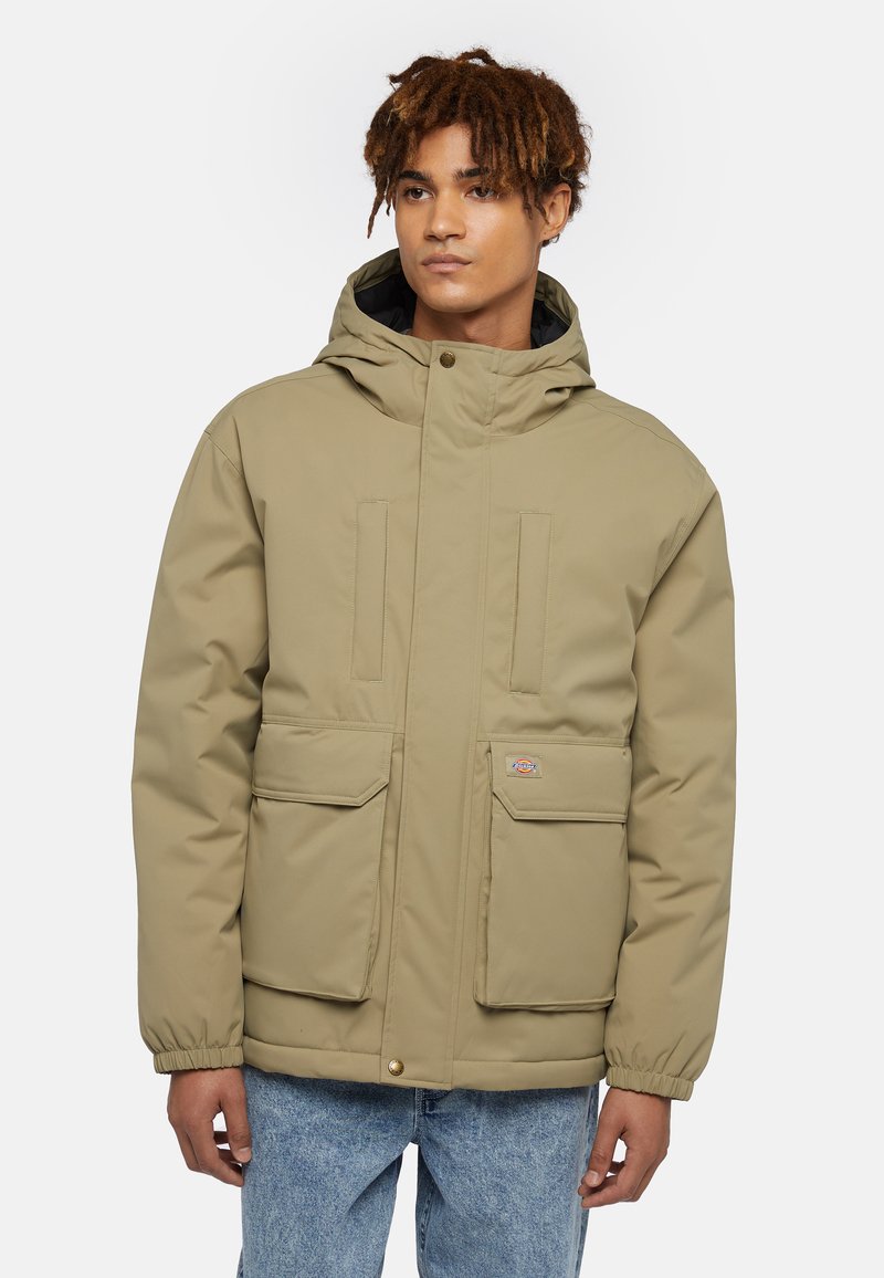 Beige vadderad jacka med huva, med två framfickor, elastiska ärmslut och en logobadge på den vänstra fickan. Slät textur.