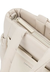 Beige laukku, jossa on pehmustettu pinta, vetoketjukiinnitys ja logotiedot. Leveät hihnat ja kompakti muotoilu helpottavat kantamista.