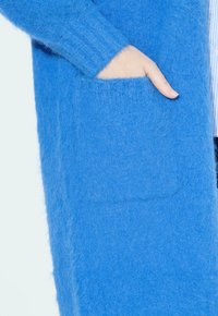 Langer blauer Cardigan aus weichem, flauschigem Material mit gerippten Bündchen und einer aufgesetzten Tasche. Besitzt eine gemütliche Textur und ein offenes Design.