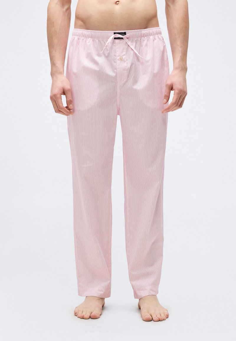 Άνδρας που φοράει ρόζ και λευκό ριγέ πιτζάμα με ελαστική μέση και κορδόνι, όρθιος ξυπόλητος μπροστά σε απλό φόντο.