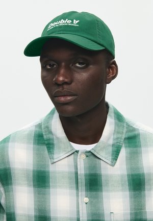 Mężczyzna w zielonej czapce z białym napisem oraz w zielono-białej kraciastej koszuli zapinanej na guziki na tle jednolitego jasnego tła.