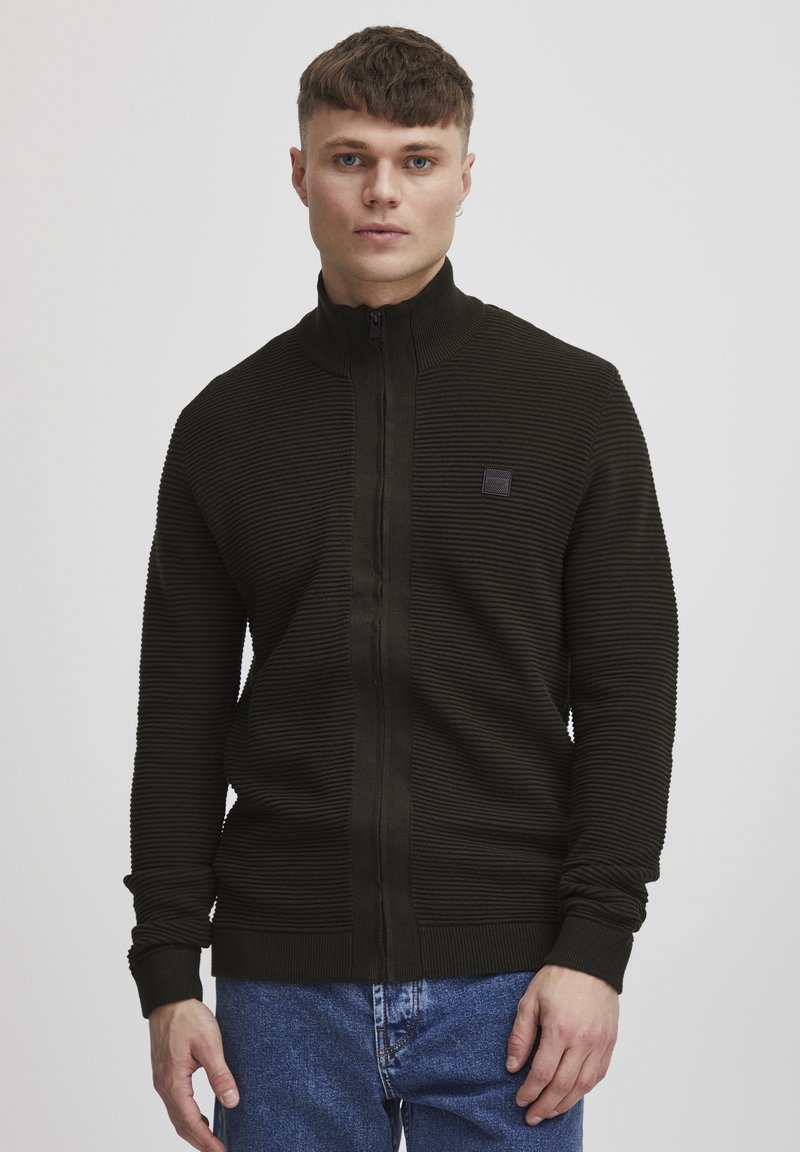 Pulover zip-up verde închis cu textură canalată și guler înalt, având o mică etichetă logo neagră pe piept, purtat peste blugi.