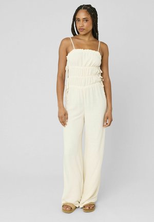 Femme aux cheveux tressés portant une combinaison sans manches de couleur crème froncée et des sandales marron clair, debout devant un fond gris uni.