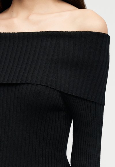 Vrouw draagt een zwarte geribbelde trui zonder schouders die sleutelbeenderen en bovenkant van de borst toont tegen een effen achtergrond.