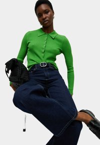 Polo shirt verde a righe con maniche lunghe e bottoni, abbinato a jeans scuri a gamba larga. Una borsa a tracolla nera intrecciata e scarpe lucide completano il look.
