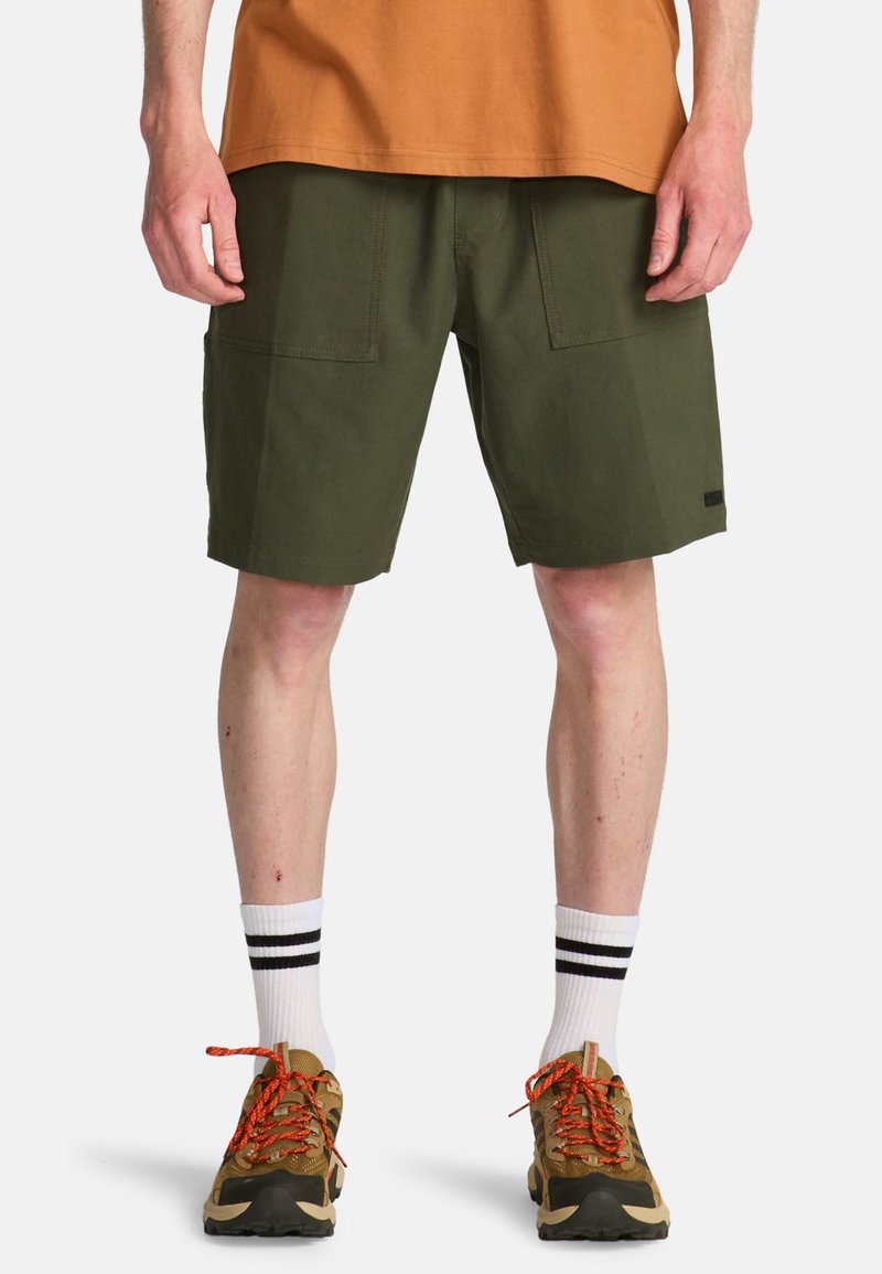 Άνδρας που φοράει ελαιί πράσινο σορτς μέχρι το γόνατο, λευκές κάλτσες με μαύρες ρίγες, καφέ πεζοπορικά παπούτσια με πορτοκαλί κορδόνια και πορτοκαλί πουκάμισο.