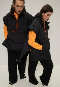 Giubbotti neri imbottiti con cerniere e dettagli arancioni, dotati di cappucci oversize, tasche a marsupio e pantaloni neri larghi.