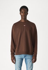 Felpa marrone con girocollo e polsini a costine, decorata con un piccolo logo rosso sul petto, abbinata a jeans di denim blu chiaro.