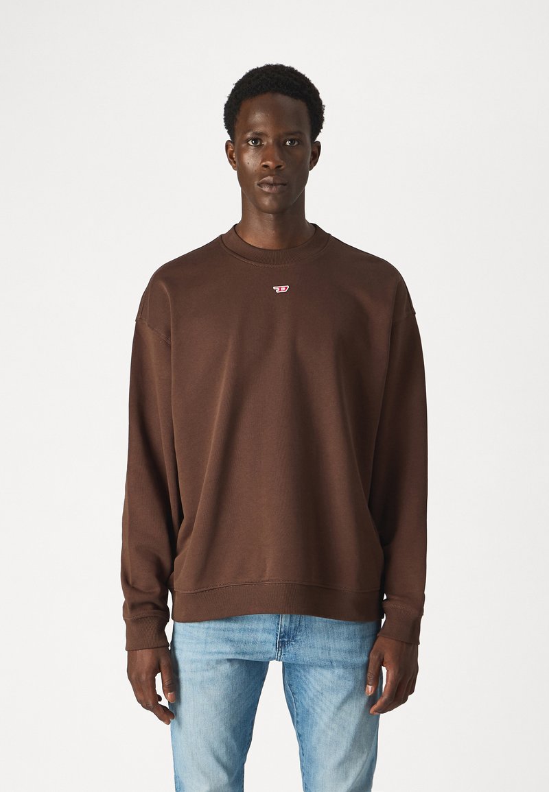 Felpa marrone con girocollo e polsini a costine, decorata con un piccolo logo rosso sul petto, abbinata a jeans di denim blu chiaro.