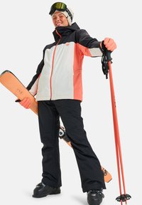 Abbigliamento da sci con una giacca impermeabile nera e corallo con cappuccio, pantaloni neri, bastoncini da sci arancioni e uno snowboard.