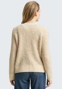 Beiger, strukturierter Pullover mit einer weichen, flauschigen Optik, gerippten Bündchen und Saum. Die Rückansicht zeigt eine lässige Passform, ergänzt durch blaue Jeans.