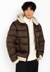 Brun pufferjakke med en hvid faux shearling-foret hætte, sorte akcenter og en lynlås foran, lagd over en cremefarvet sweater.