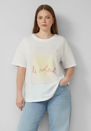 Junge Frau mit glatten braunen Haaren, die ein weißes oversized T-Shirt mit gelber Sonne und dem Schriftzug „le soleil“ trägt, kombiniert mit hellblauen Jeans.