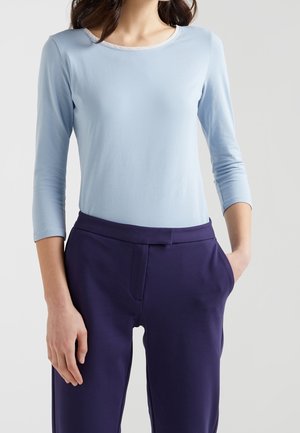 Femme portant un haut bleu clair à manches longues avec un col rond et un pantalon bleu marine foncé, debout avec une main dans la poche.