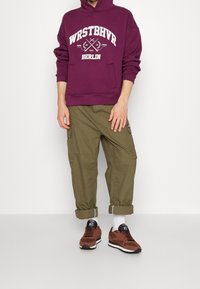 Paarse hoodie met witte grafische tekst, olijfgroene cargo pants met opgerolde boorden, en bruine sneakers met zwarte accenten.