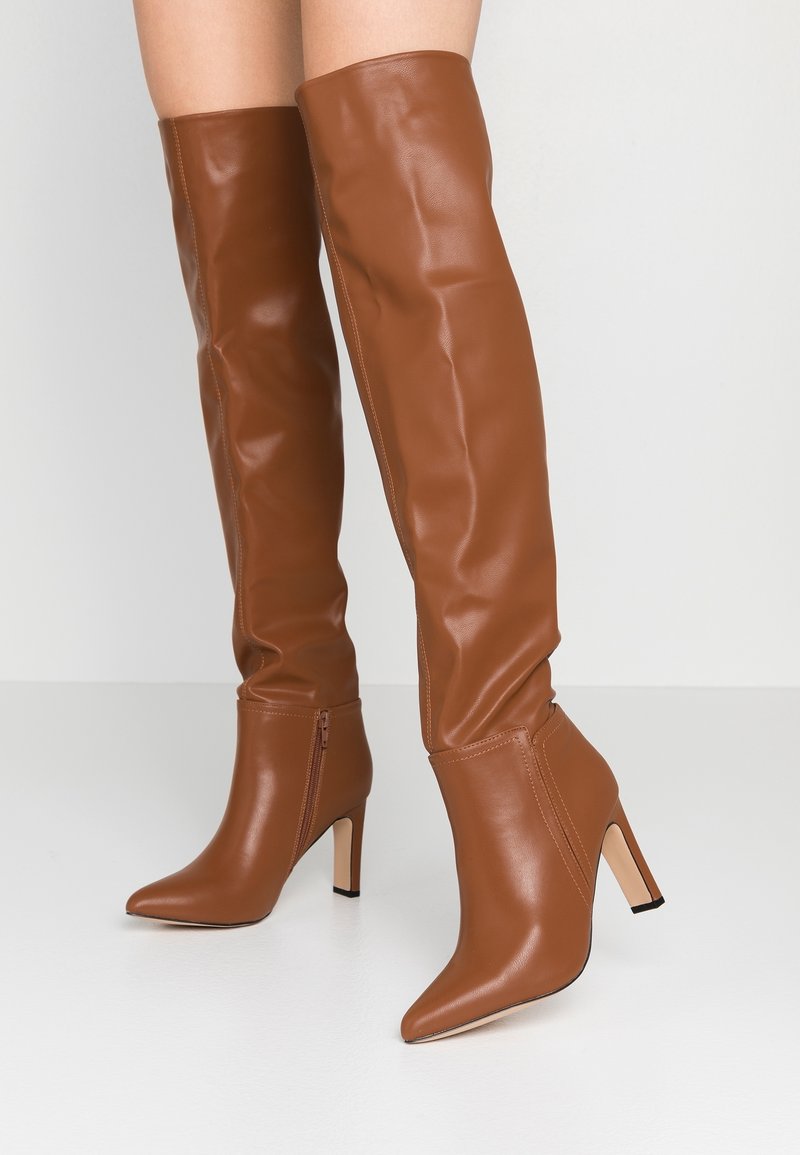 Botas largas marrones con tacón en cuero liso. Presentan punta afilada y cremallera lateral. El diseño incluye una forma ajustada que se extiende por encima de la rodilla.