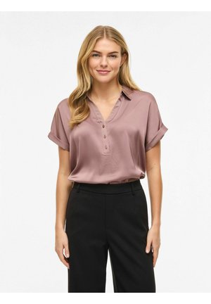 Femme blonde debout, portant un chemisier mauve à manches courtes avec des boutons et un pantalon noir taille haute sur un fond blanc uni.