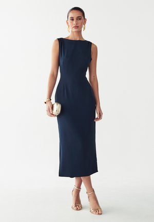 Vrouw draagt een mouwloze navy midi-jurk, gouden oorbellen, armband, houdt een gouden clutch vast en gouden sandalen met hoge hakken en enkelbandje.