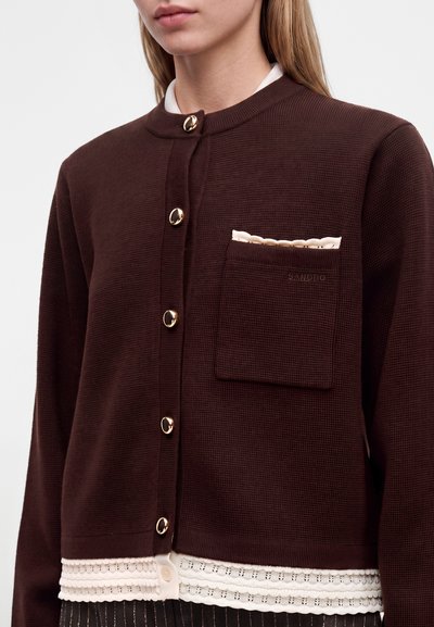Femme portant un cardigan en tricot marron foncé avec des boutons dorés et une bordure en dentelle crème à l'ourlet, superposé à un haut blanc.