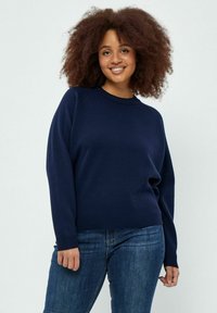 Marineblaues Rundhals-Sweatshirt mit Raglanärmeln, weicher Textur und gerippten Bündchen. Getragen mit dunkelblauen Jeans.