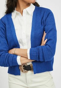 Modrý pletený cardigan s výstřihem do V, žebrovanými manžetami a lemem. Nošený přes bílou košili s límečkem, doplněný velkou ozdobnou sponou na opasku.