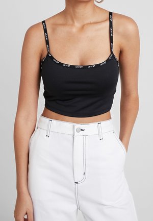Woman wearing a black cropped tank top with Levi's logo straps and high-waisted white pants with contrast stitching and front pockets.