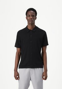 Black short-sleeve polo shirt with a collar, featuring a button-up front and a textured fabric. Worn with light gray pants.