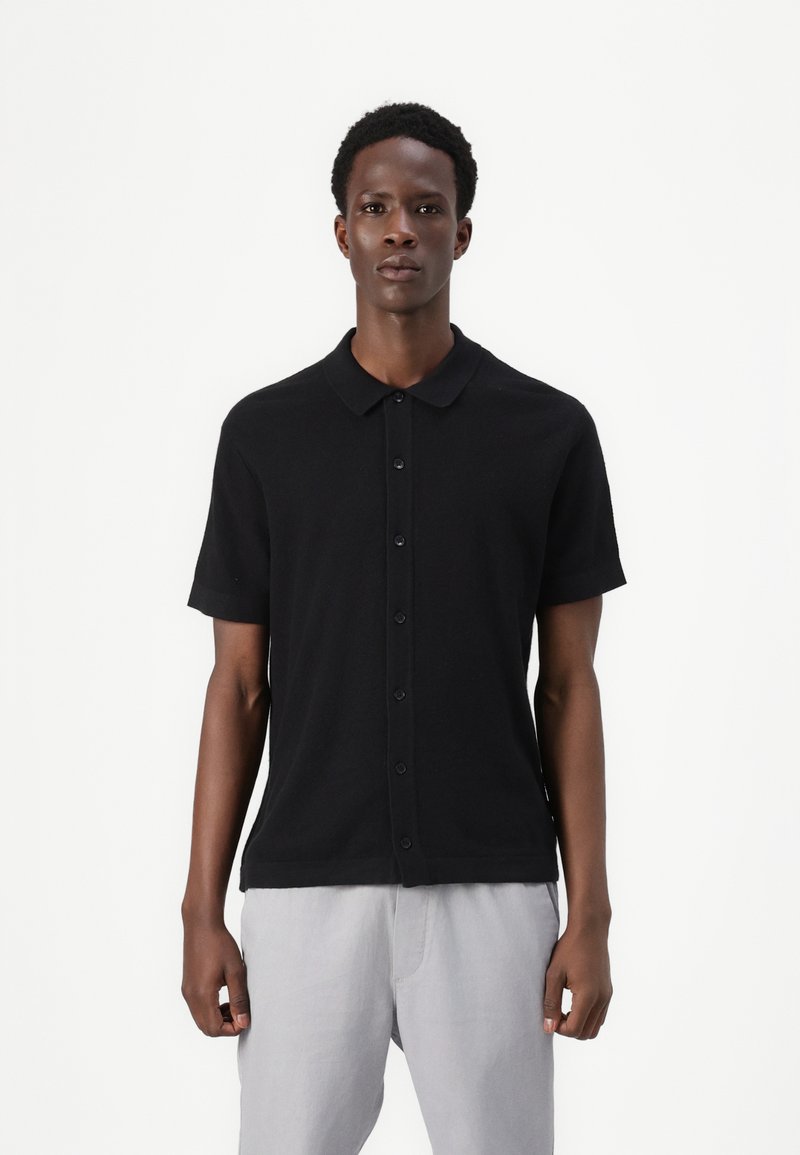 Black short-sleeve polo shirt with a collar, featuring a button-up front and a textured fabric. Worn with light gray pants.