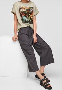 Jasnozielony bawełniany t-shirt z kolorowym tropikalnym nadrukiem i napisem "SUNSET LOVE", w połączeniu z szerokimi szarymi lnianymi spodniami i czarnymi sandałami.