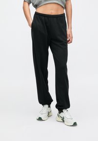 Μαύρο παντελόνι φόρμας με χαλαρή εφαρμογή και ελαστική μέση, με πλαϊνές τσέπες και μανσέτες στους αστραγάλους, σε συνδυασμό με λευκά και πράσινα αθλητικά παπούτσια.
