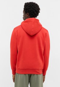 Mężczyzna z krótkimi kręconymi włosami, ubrany w czerwoną bluzę z kapturem i zielone spodnie, stoi tyłem na jednolitym tle.