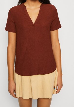 Femme portant un haut rouille à manches courtes avec un col en V et une jupe plissée beige avec une petite figure brodée près de l'ourlet.