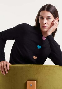 Schwarzer Pullover mit vier filzartigen Herz-Applikationen in Pink, Blau, Braun und Beige. Modell sitzt auf einem grün gepolsterten Stuhl.
